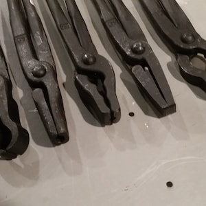May include: Six black metal blacksmith tongs with different jaw shapes and sizes.