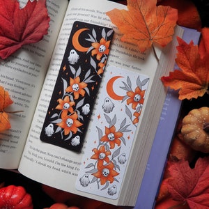 May include: Two Halloween-themed bookmarks with a black and white design featuring a crescent moon, ghosts, and orange flowers with skulls. The bookmarks are on a book with fall leaves and pumpkins.
