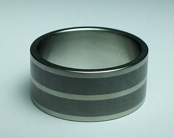 May include: A wide, silver-colored ring with two dark gray bands. The ring has a simple, modern design and is likely made of metal. The bands are evenly spaced around the ring's circumference.