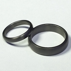 May include: Two dark gray rings are displayed against a white background. One ring is thinner and more delicate, while the other is wider with a rounded band. The rings appear to be made of metal.