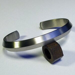 May include: A silver-colored cuff bracelet with a brushed metal finish. The bracelet has a simple, modern design with a slightly angular shape. The open ends allow for easy wear. The bracelet is resting on a rusty metal nut.