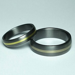 May include: Two dark gray rings with a gold-colored band. The rings are made of a dark gray metal with a brushed finish. One ring is smaller than the other. The gold band is centered on each ring.