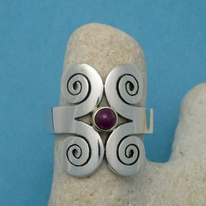 May include: A silver ring with a central, round, dark red gemstone. The ring features four swirling spiral designs and a gold-colored setting around the gemstone. The ring is displayed on a light-colored, textured surface against a blue background.