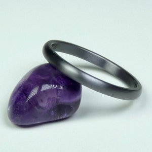 May include: A dark gray bangle bracelet rests on a polished, deep purple amethyst stone. The bracelet has a smooth, matte finish and a simple, minimalist design. The amethyst is oval-shaped and reflects light.