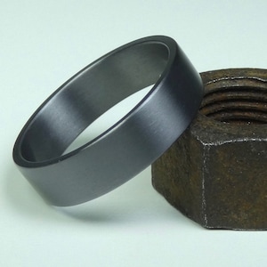 May include: A dark gray ring with a flat top and rounded edges. The ring is next to a rusty, hexagonal nut. The ring has a smooth, matte finish. The ring is likely made of metal.