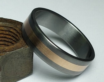 May include: A close-up of a ring with a dark gray exterior and a central band of rose gold. The ring is resting on a rusty metal nut, set against a white background. The ring's design is simple and modern.