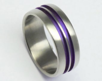 May include: A silver-colored ring with two parallel purple inlays. The ring has a brushed metal finish and a rounded, rectangular shape. The purple lines add a pop of color to the otherwise neutral design.