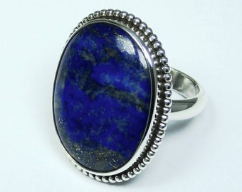 May include: A sterling silver ring featuring a large, oval-shaped lapis lazuli stone. The deep blue stone has streaks of gold and is set in a beaded bezel. The ring's band is smooth and polished.