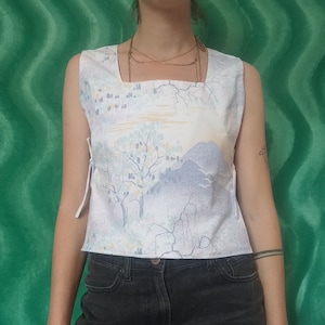 Könnte beinhalten: Ärmelloses Oberteil mit quadratischem Ausschnitt und Landschaftsdruck in Pastellfarben, mit Bäumen und Bergen. Das Oberteil hat weiße Bindebänder an den Seiten und wird mit schwarzen Jeansshorts getragen.