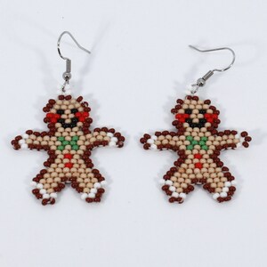 May include: A pair of festive gingerbread man earrings. Each earring is crafted with small, tan, brown, red, green, and white beads, forming a gingerbread man shape. The earrings have silver-colored hooks.