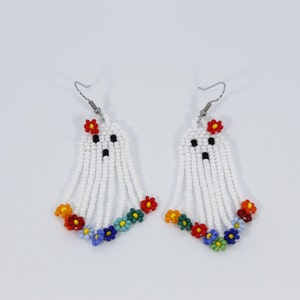 May include: A pair of white beaded ghost earrings with colourful flower accents. The earrings are made with small white beads and feature a ghost face with black eyes and a black mouth. Each ghost has a red flower on top and a row of colourful flowers at the bottom.