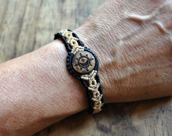 Puede incluir: Pulsera de macramé con una pieza central de madera con un diseño de sol y luna. La pulsera es negra y beige, con un patrón tejido. La pieza central de madera es redonda y tiene un símbolo de sol y luna tallado.