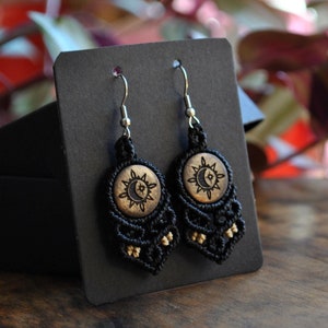 May include: A pair of black macrame earrings with a sun and moon design on a wooden button. The earrings have silver-colored hooks and small gold-colored beads. The earrings are displayed on a black card.