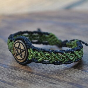 May include: A macrame bracelet featuring a central circular wooden charm engraved with a black pentagram. The band is constructed from interwoven black and olive green cord, showcasing a detailed knotted pattern and adjustable cord ends.