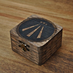 Peut inclure: Petite boîte carrée en bois teinté foncé. Le couvercle présente un motif circulaire avec trois lignes rayonnantes et une bordure décorative. La boîte a un fermoir et des charnières en métal, posée sur une surface en bois.