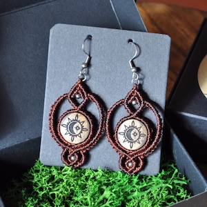 Puede incluir: Un par de pendientes de macramé marrones con un diseño de sol y luna. Los pendientes cuelgan de ganchos plateados y se muestran en una tarjeta gris.