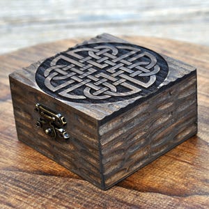 Puede incluir: Una caja cuadrada de madera, teñida de color marrón oscuro, con un diseño de nudo celta tallado en la tapa. La caja tiene un exterior texturizado y tallado y se asegura con un pequeño cierre de estilo antiguo. Dimensiones: aproximadamente 10 cm x 10 cm.
