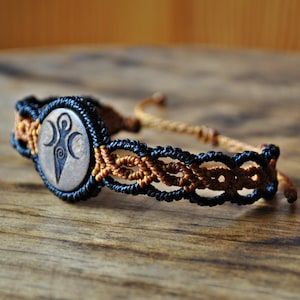 May include: A macrame bracelet with a central wooden bead, engraved with a symbol. The bracelet is made with black and brown cords, displaying intricate knotting. The adjustable closure is made of a tan cord. The bracelet is approximately 18 cm in length.