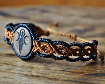 Puede incluir: Pulsera de macramé hecha a mano con una cuenta central de madera con un símbolo grabado. La pulsera está hecha con cordones negros y marrones, mostrando intrincados patrones de nudos. El cierre ajustable es de cordón marrón, aproximadamente 18 cm.