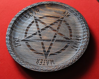 May include: A round, wooden tray with a carved pentagram design. The tray is stained in shades of gray and brown, with the words "Earth, Air, Fire, Water, Spirit" carved around the edges. The tray has a decorative border and sits on a red surface.