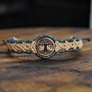 Puede incluir: Pulsera de macramé con un colgante de madera con el árbol de la vida. La pulsera tiene un diseño tejido con hilos beige y grises. El colgante del árbol de la vida es circular y está hecho de madera, con un grabado detallado del árbol.