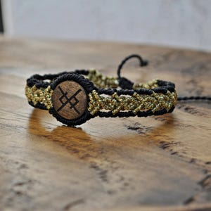 Könnte beinhalten: Ein Makramee-Armband mit einer zentralen Holzperle mit einem eingravierten Symbol. Das Armband kombiniert schwarze und goldfarbene Fäden in einem gewebten Muster. Es hat einen verstellbaren Verschluss aus schwarzem Kordel. Dieses handgefertigte Accessoire ist für den Alltag geeignet.