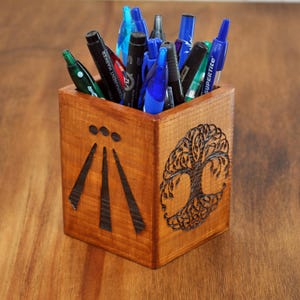 Könnte beinhalten: Holzstiftbehälter mit einem Lebensbaum-Design und einem schwarz-weißen Symbol auf der Vorderseite. Der Behälter ist mit verschiedenen Stiften und Bleistiften gefüllt.