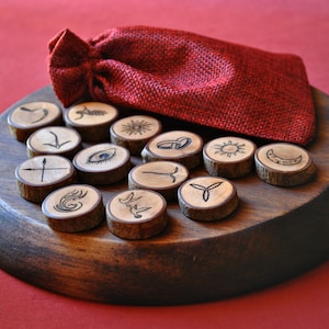May include: A set of wooden rune stones with etched symbols, displayed on a wooden tray. A red burlap drawstring bag is nestled behind the stones. The runes are various shapes and designs, with a natural wood grain.