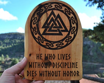 May include: Wooden plaque featuring a carved black Valknut symbol within a circular knot design. The plaque has the quote, "HE WHO LIVES WITHOUT DISCIPLINE DIES WITHOUT HONOR" etched in black. The wood has a natural grain and a light brown colour.