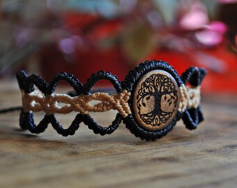 Puede incluir: Pulsera de macramé con un centro de madera con un diseño de árbol de la vida. La pulsera tiene un patrón tejido en negro y beige. El centro de madera es redondo y tiene un diseño de árbol marrón oscuro. La pulsera está sobre una superficie de madera.