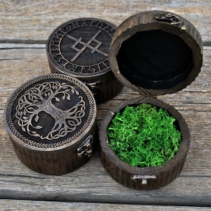 May include: Three round wooden boxes with intricate carvings. One box features a tree of life design, another displays runes, and the third is open, revealing green moss inside. The boxes have a dark brown finish and are set on a wooden surface.