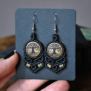 Puede incluir: Pendientes de macramé negro con un dije de árbol de la vida de madera. Los pendientes cuelgan de ganchos plateados.