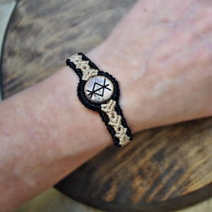 Könnte beinhalten: Makramee-Armband mit einem zentralen kreisförmigen Anhänger mit einem schwarzen Runensymbol. Das Armband ist aus schwarzen und beigefarbenen Makramee-Knoten gefertigt. Der Anhänger hat eine silberfarbene Metalloberfläche. Das Armband wird am Handgelenk getragen.
