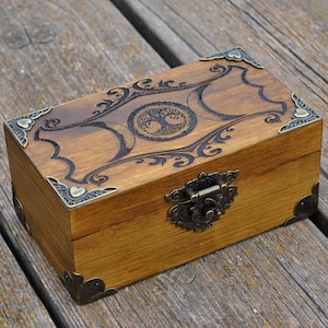 May include: A rectangular wooden box with a carved tree of life design on the lid. The box has decorative metal corner pieces and a clasp. The wood has a warm, natural tone, and the carvings are dark.