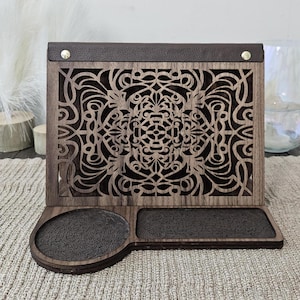May include: A wooden phone stand with a decorative laser-cut design and two slots for holding items. The stand has a brown leather top and a dark brown finish.