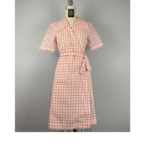 Pode incluir: Vestido vintage envelope com padrão xadrez rosa e branco. O vestido apresenta mangas curtas, decote com gola, cinto a condizer e um bolso. O vestido é exibido num manequim.
