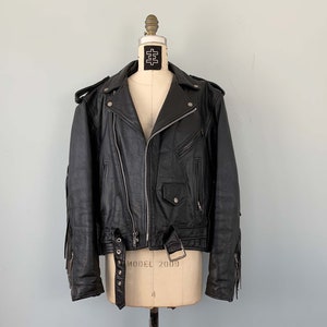 Puede incluir: Una chaqueta de cuero negra para moto con detalles de flecos en las mangas. La chaqueta tiene cierre de cremallera, cinturón en la cintura y varios bolsillos.