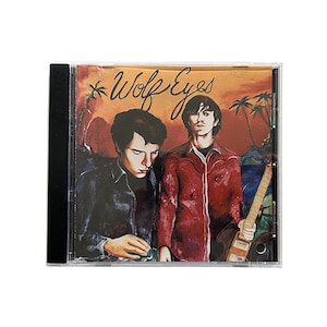 Op de afbeelding: CD-hoesje met de handgeschreven tekst "Wolf Eyes". De cover toont twee mannen, een in een zwart overhemd en de ander in een rood overhemd met een gitaar. De achtergrond toont een zonsondergang en palmbomen.