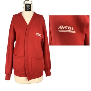 May include: Red cardigan sweater with a button-up front and two pockets. The sweater has the word "AVON" embroidered on the front in white with a star and three stripes.