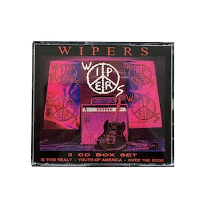 Op de afbeelding: Een cd-boxset van de band Wipers. De hoes heeft een paars en roze ontwerp met de naam en het logo van de band. De set bevat drie cd's: "Is This Real?", "Youth of America" en "Over the Edge". Een gitaar en een versterker zijn ook zichtbaar.