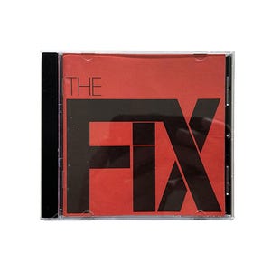 Op de afbeelding: Een cd-hoesje met een rode cover met de zwarte tekst "THE" boven "FIX". De cd-hoes is transparant met een zwarte rug. De tekst is in een vetgedrukte, schreefloze lettertype.