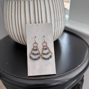 May include: Copper-colored earrings with a layered, triangular design. Each layer is adorned with small, round beads in shades of green and red. The earrings are displayed on a light gray card, set against a blurred background.