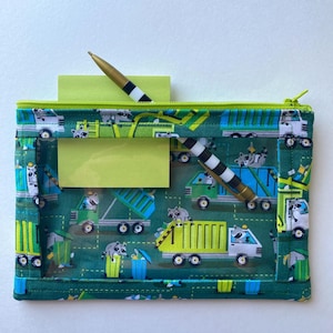 Könnte beinhalten: Grünes Federmäppchen mit einem Muster aus Waschbären und Lastwagen. Das Etui hat einen neongrünen Reißverschluss und Rand. Zwei Stifte mit schwarz-weißen Streifen befinden sich in dem Etui, zusammen mit einem gelben Haftzettel.