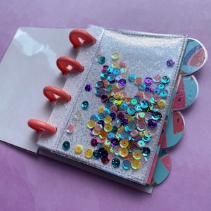 May include: A small notebook with a clear, glitter-filled cover and colourful sequins. It features peach-coloured binding rings and watermelon-themed divider tabs. The notebook is open, showing blank white pages.