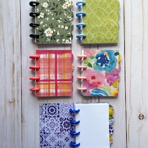 May include: Six mini notebooks with decorative covers and colorful binding rings. The covers feature floral, plaid, and patterned designs in various colors. The notebooks are arranged on a light wood-grain surface.