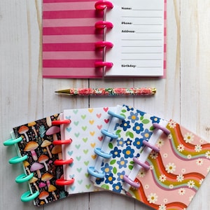 May include: A collection of mini notebooks with various designs, including mushrooms, hearts, and flowers, with colorful disc binding. A floral pen and a pink striped notebook with a contact information page are also present. The notebooks are arranged on a light wood surface.