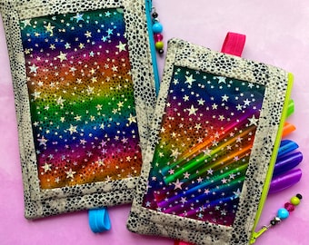 May include: Two zippered pouches with clear vinyl windows. The pouches are made of a beige fabric with a black and white speckled pattern. The pouch on the left has a rainbow holographic background with white stars. The pouch on the right has a rainbow holographic background with white stars and is filled with colorful pens.