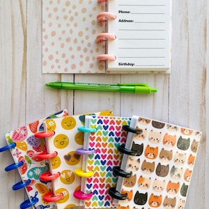 May include: Mini notebooks with colorful covers and ring binding. Designs include butterflies, smiley faces, hearts, and cats. One notebook is open, displaying lines for name, phone, address, and birthday. A green pen rests nearby.