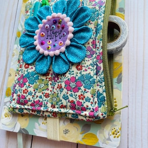 May include: A fabric notebook cover featuring a floral pattern in pink, blue, and green, with a large teal fabric flower appliqué centered on the front. The flower has a lavender felt center embroidered with the word Hope and colorful sequins.