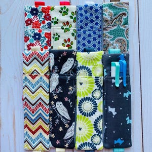 May include: A collection of fabric pen holders in various patterns. Designs include floral, paw prints, geometric, paisley, owl, and floral patterns. Each holder has a small loop and a contrasting ribbon tab. Some holders contain pens and erasers.